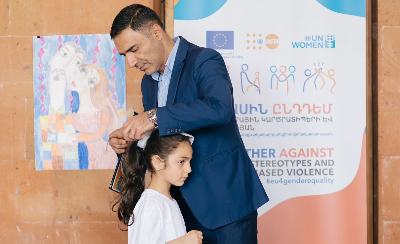 Image of a father assisting his daughter in putting on her cap in front of a banner.