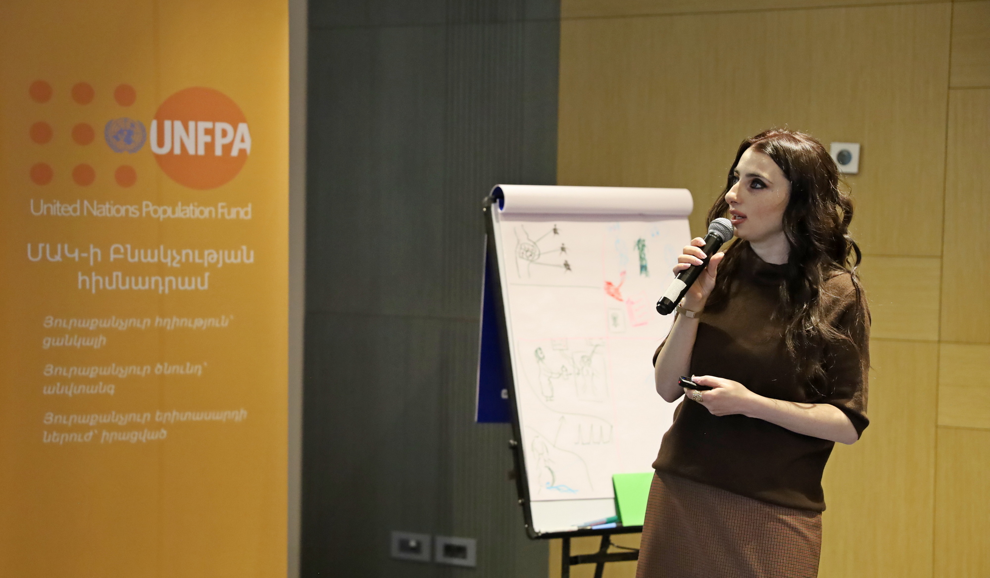 The image depicts a woman speaking into a microphone, in the background, the banner of the United Nations Population Fund (UNFPA).