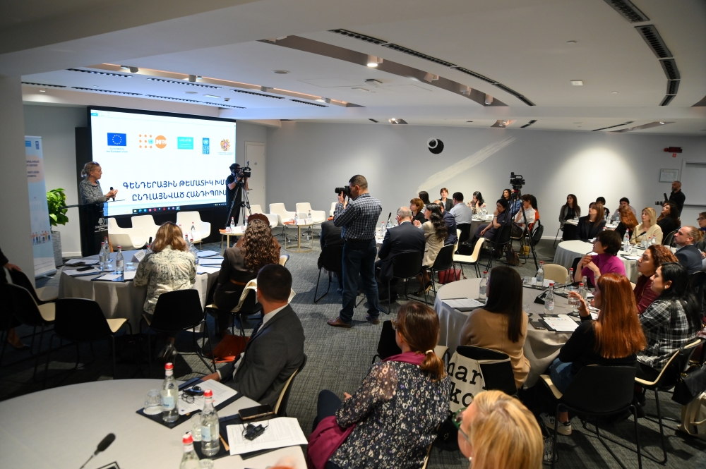 Attendees seated at a conference listening to a speaker at a podium, with cameras filming and event logos of various organizations displayed in the background.