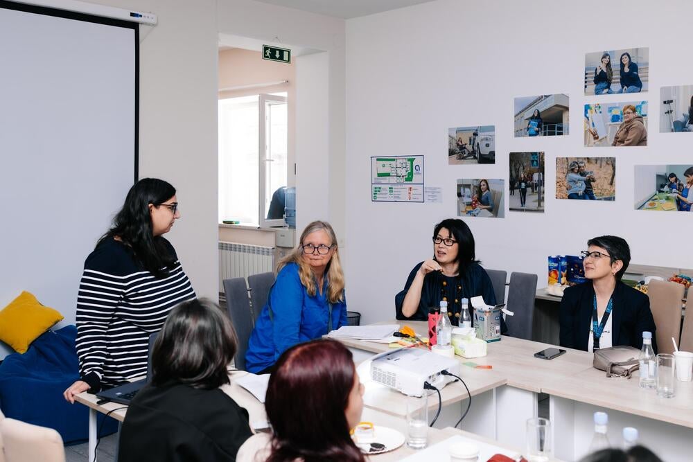 A group of women are gathered in a bright, modern meeting room. One woman is standing and speaking while others sit around a table, listening attentively and engaging in discussion. 