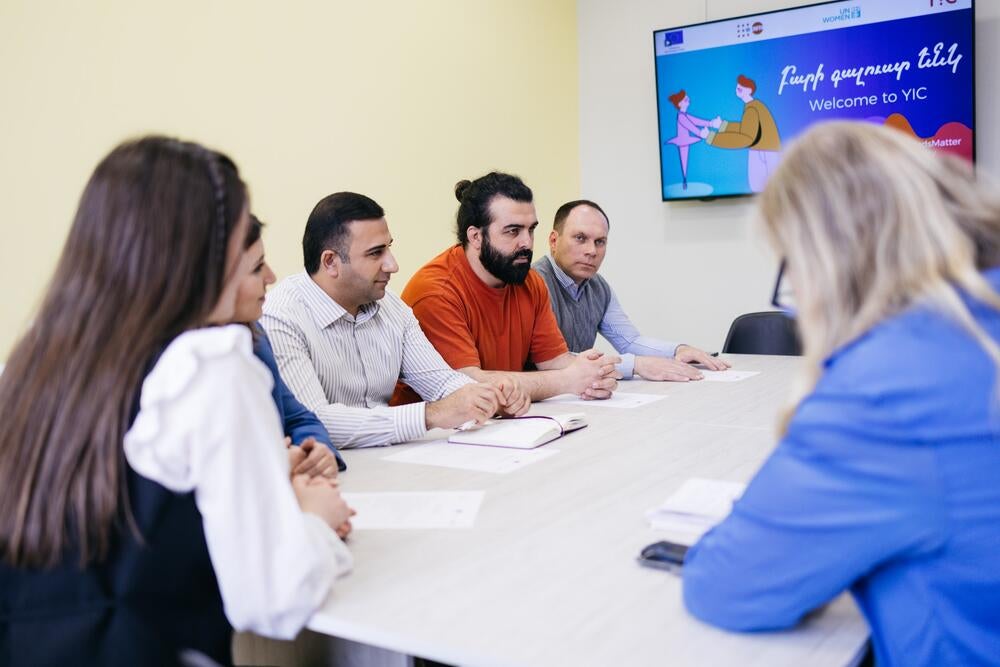 A group of professionals in a meeting room with one person presenting at a table, and a digital screen in the background.
