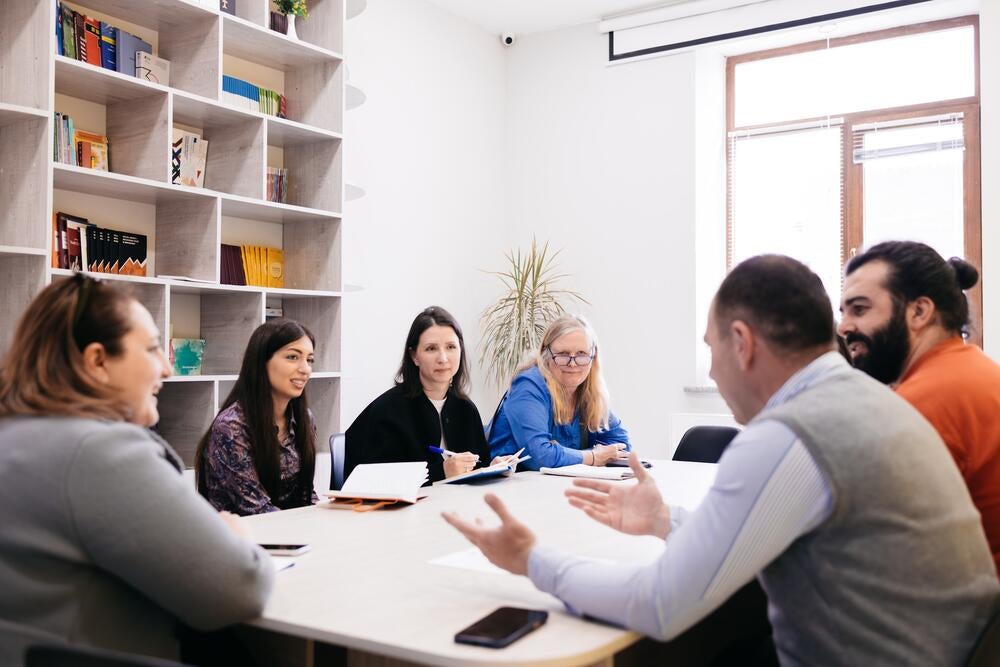 Six people—four women and two men—sit around a table in a bright room, engaged in a lively discussion. They are smiling and listening attentively to one another.