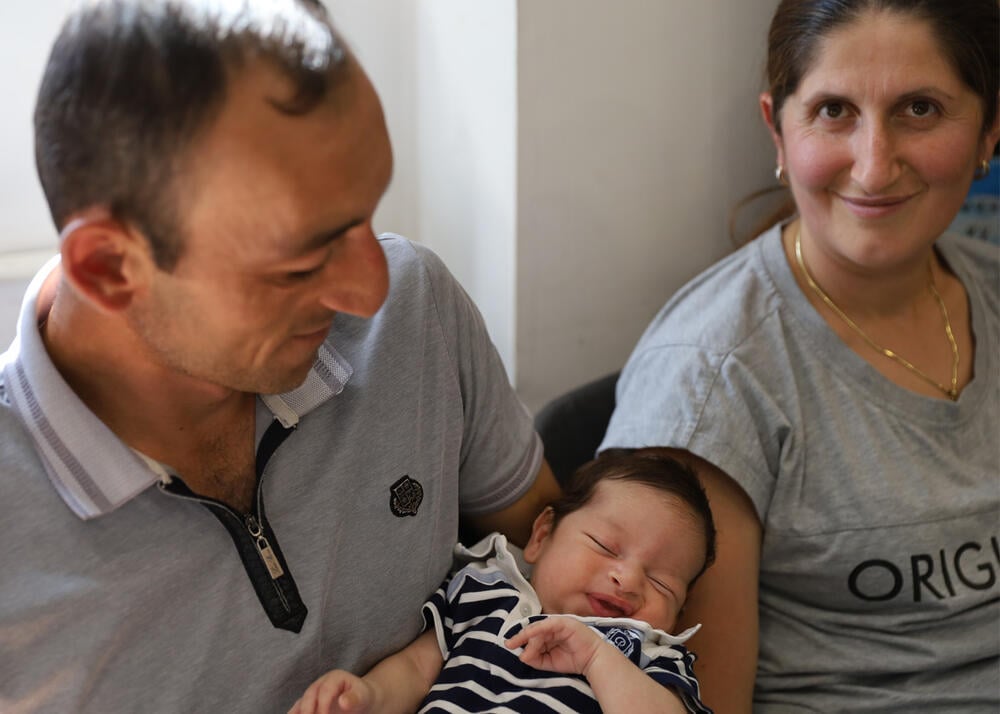 Image of a young Chichyan family with their newborn child. The father is holding the baby and smiling at him, while the mother is looking at the camera.