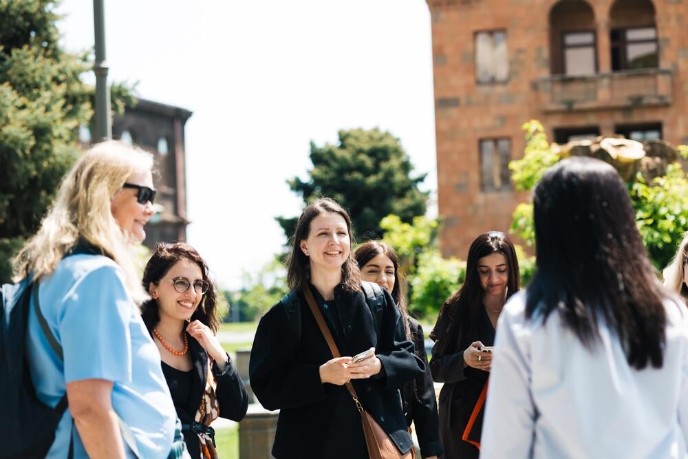 A group of individuals engaged in conversation outdoors on a sunny day, with trees and a historical building in the background.