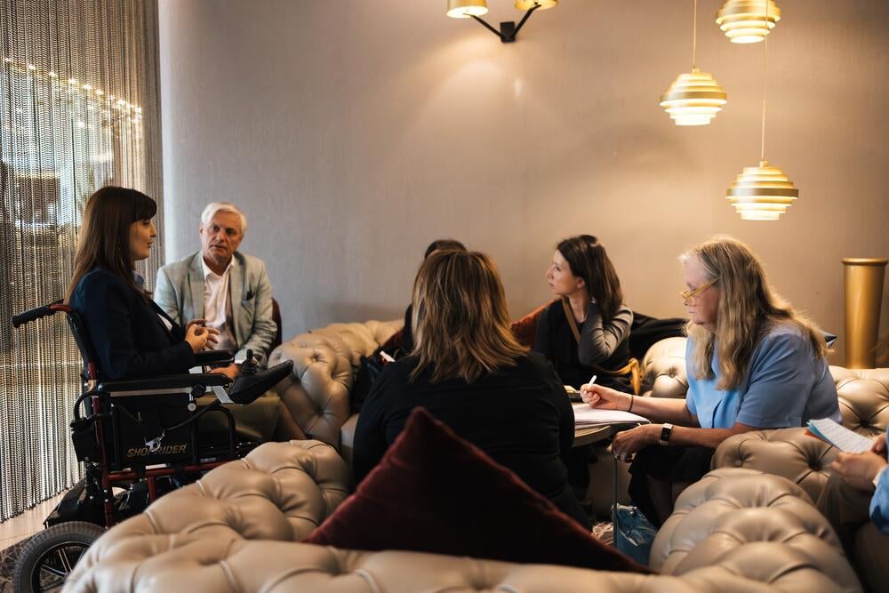 A group of five people, one using a wheelchair, engaging in a meeting in a stylish room with pendant lights and a tufted sofa.
