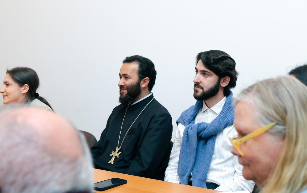  A priest, a man in a white shirt with a blue sweater, and a woman sit side by side at a table, attentively listening during a group discussion. Warm expressions and focused attention suggest a thoughtful, inclusive atmosphere.