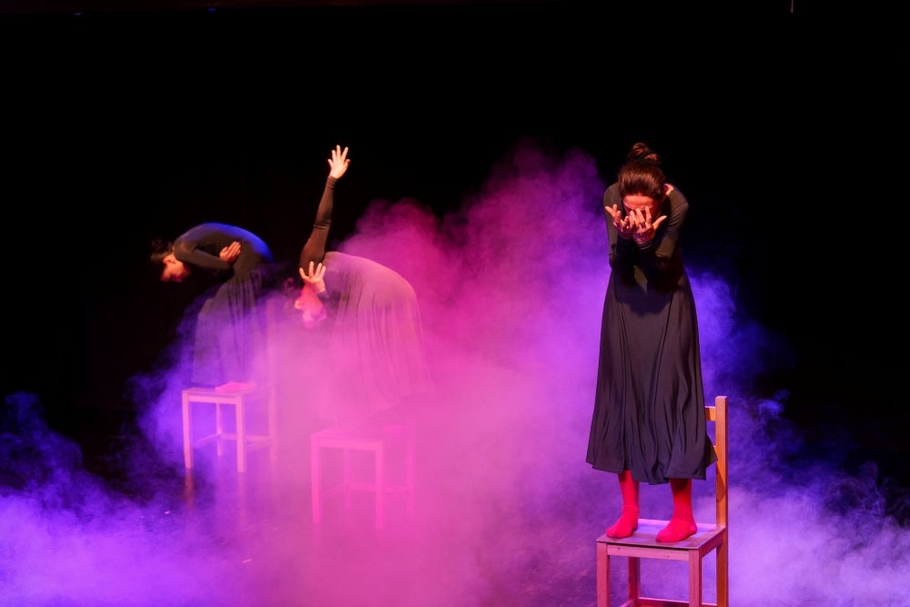 A theatrical performance featuring three individuals on stage with dramatic blue and purple lighting and mist effects. One person is standing on a stool, holding their head, while the others seems to be reaching out expressively from behind a chair.