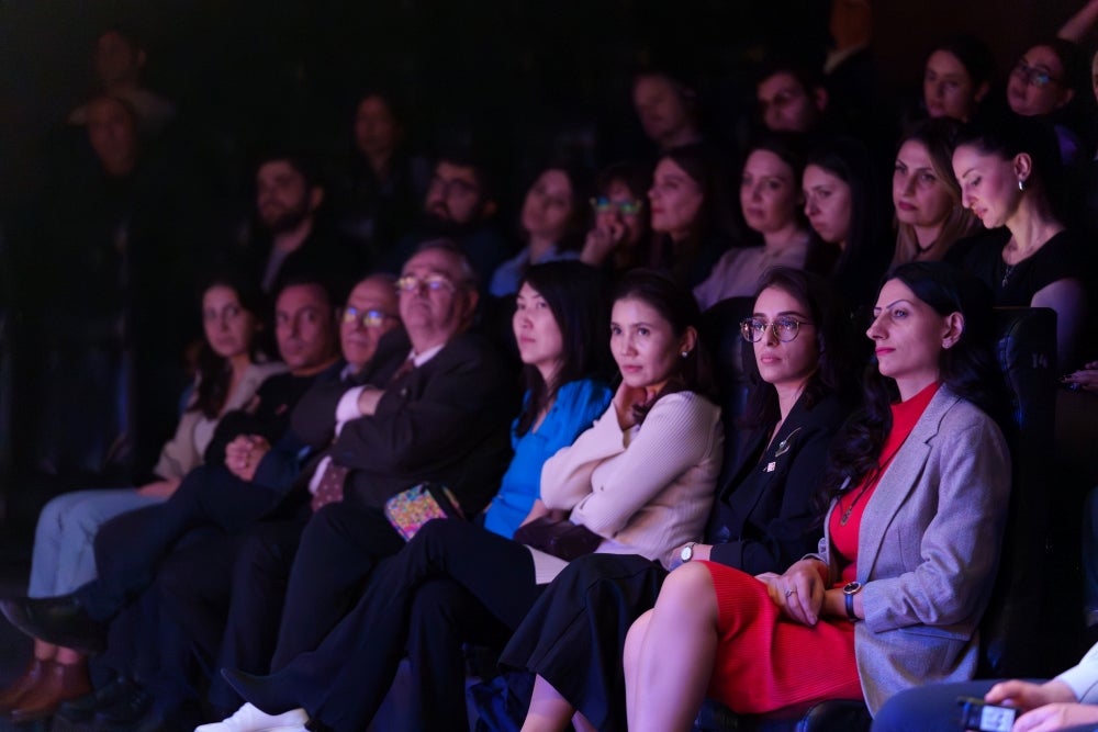 Audience members with focused expressions watching a presentation or performance in a dimly lit auditorium.