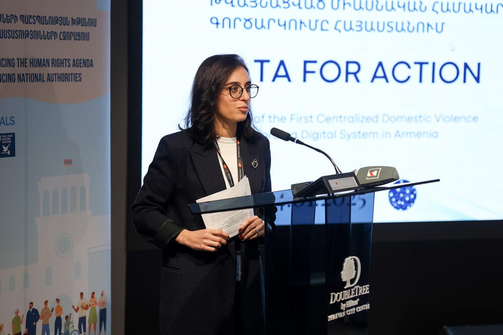 A woman stands at a podium with a microphone, presenting at the launch event of the first centralized domestic violence case management digital system in Armenia. Behind the speaker, a banner displays the event's title "Data for Action" along with various logos including UNFPA and the European Union.