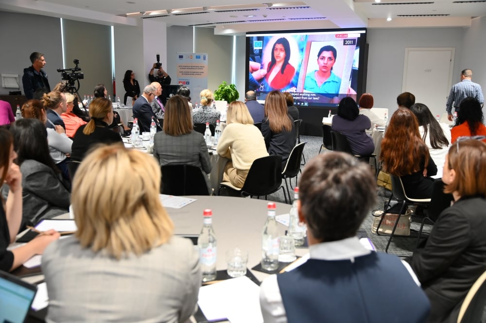 Attendees at a conference watching a presentation with photos of women displayed on a large screen, in a well-lit room equipped with modern technology.