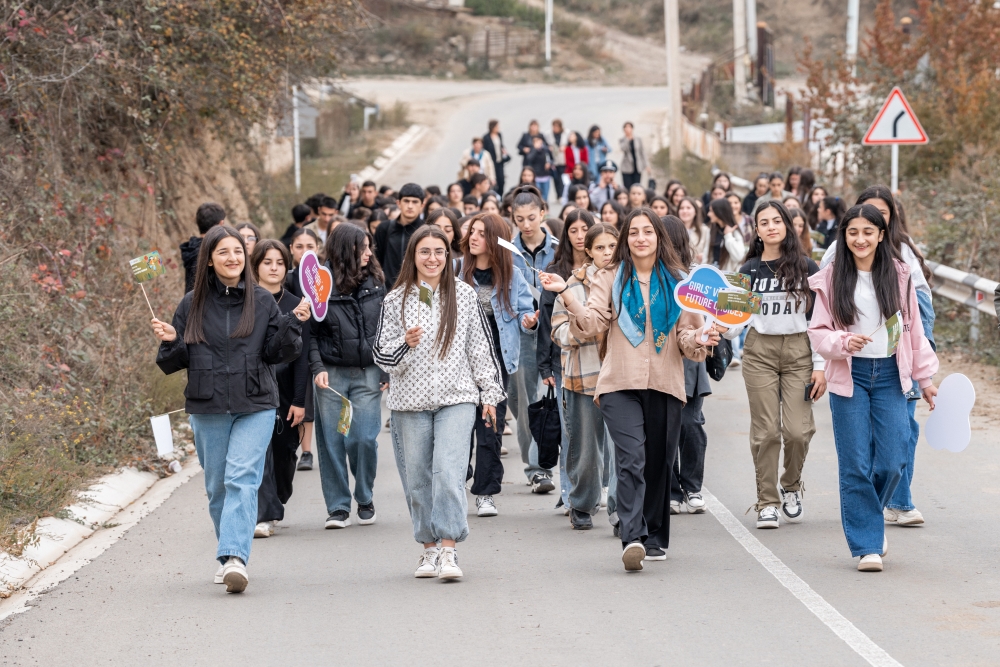 A group of young girls and boys walking along a road, some holding flags and stands, in a lively outdoor setting.