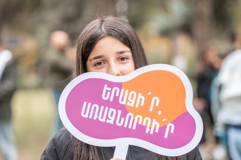 A girl holding a colorful sign with Armenian text, reading 'Dream, Lead' at an outdoor event.