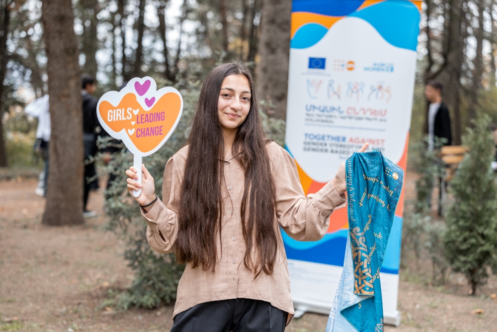 A person holding a sign that reads "GIRLS LEADING CHANGE" and displaying a blue scarf, standing in front of “EU 4 Gender Equality: Together against gender stereotypes and gender-based violence” programme banner.