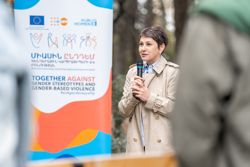 Person standing next to the “EU 4 Gender Equality: Together against gender stereotypes and gender-based violence” programme banner, holding a microphone and speaking at an outdoor event.