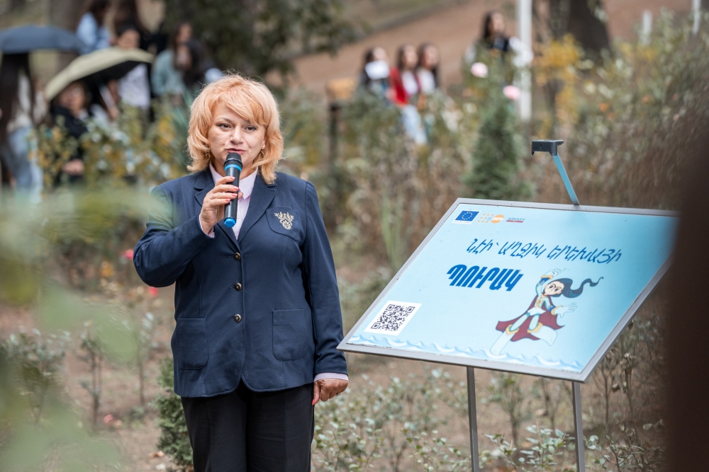 A person in a blue blazer speaks into a microphone at an outdoor event next to a sign, which reads "Girl Child NE's Park"