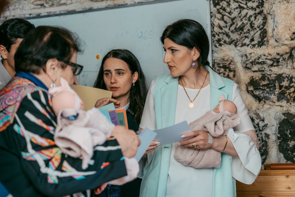 Three women are in discussion, holding colorful papers and lifelike baby dolls. One woman in a light vest holds a swaddled doll while listening intently. They appear to be participating in a training or educational session in a room with a stone wall and whiteboard in the background.