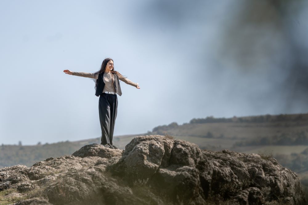 A girl stands confidently on a rocky cliff with her arms stretched wide, embracing the open air. 