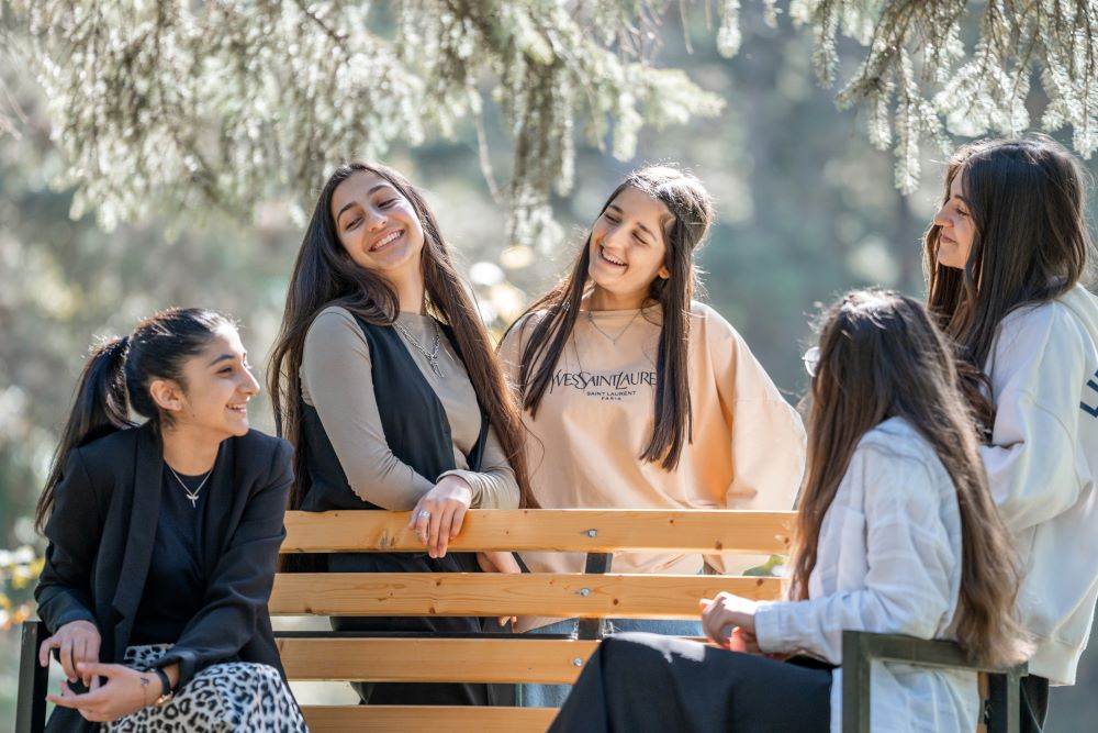 A group of five young girls laughing and enjoying a conversation on a park bench surrounded by trees.