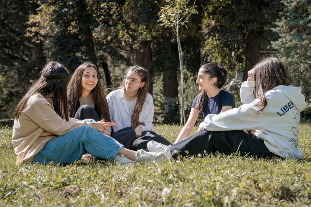 Five girls sitting on the grass in a park, engaged in a lively conversation and enjoying a sunny day.