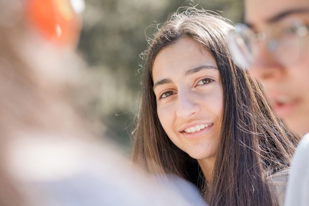 Close-up of a smiling girl outdoors in a sunny setting, partly obscured by another person in the foreground.