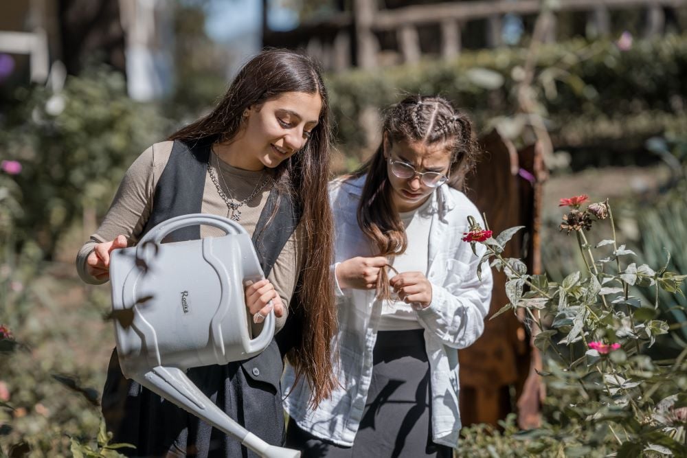 Two girls examining plants in a garden, one holding a watering can.