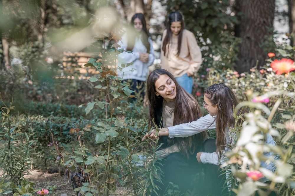 Two individuals smiling and examining roses in a lush garden, with two others in the background engaged in conversation.