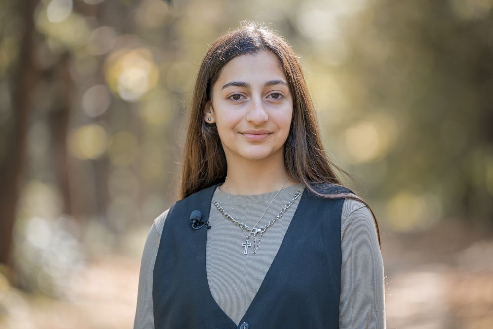 Portrait of a smiling girl wearing a grey vest and a cross necklace, standing in a sunlit wooded area.