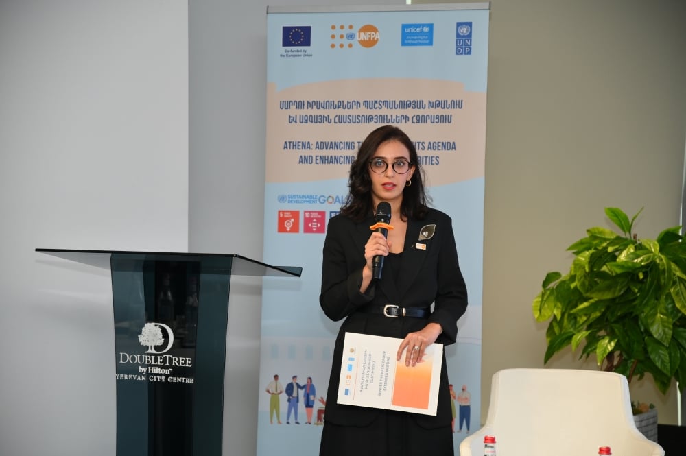 Lusine Sargsyan, UNFPA Armenia Head of Office standing at a podium with a microphone, holding documents, with banners displaying logos from the EU, UNICEF,UNFPA and UNDP in the background