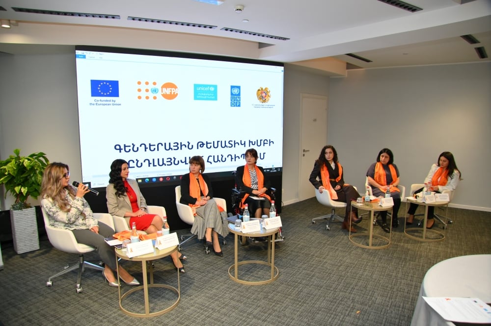 Panel discussion at an event with logos of European Union, UNFPA, UNICEF, and UN Women displayed on a screen behind the speakers who are seated in a row, wearing orange scarves. Several audience members are seated at tables facing the panel.