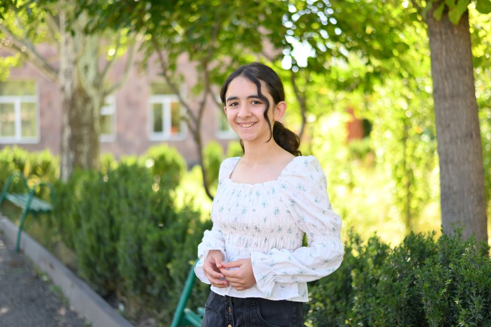 A girl smiling in a garden with trees and a building in the background.