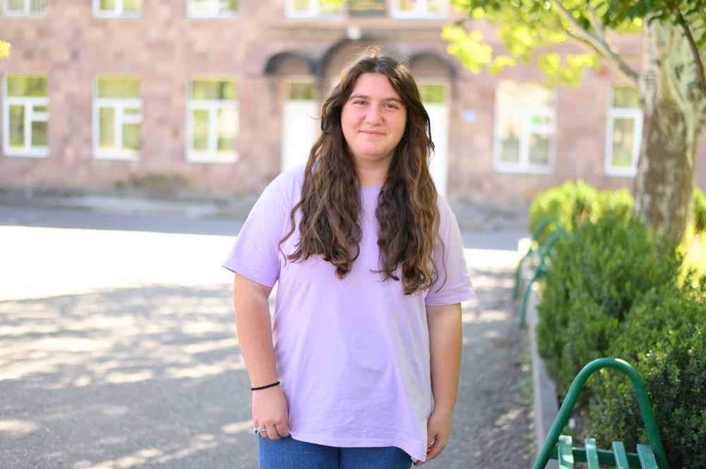 A girl in a purple t-shirt standing in front of a school building with trees. 