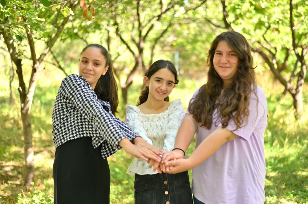 Three girls standing outdoors, holding hands with smiles, surrounded by trees. 