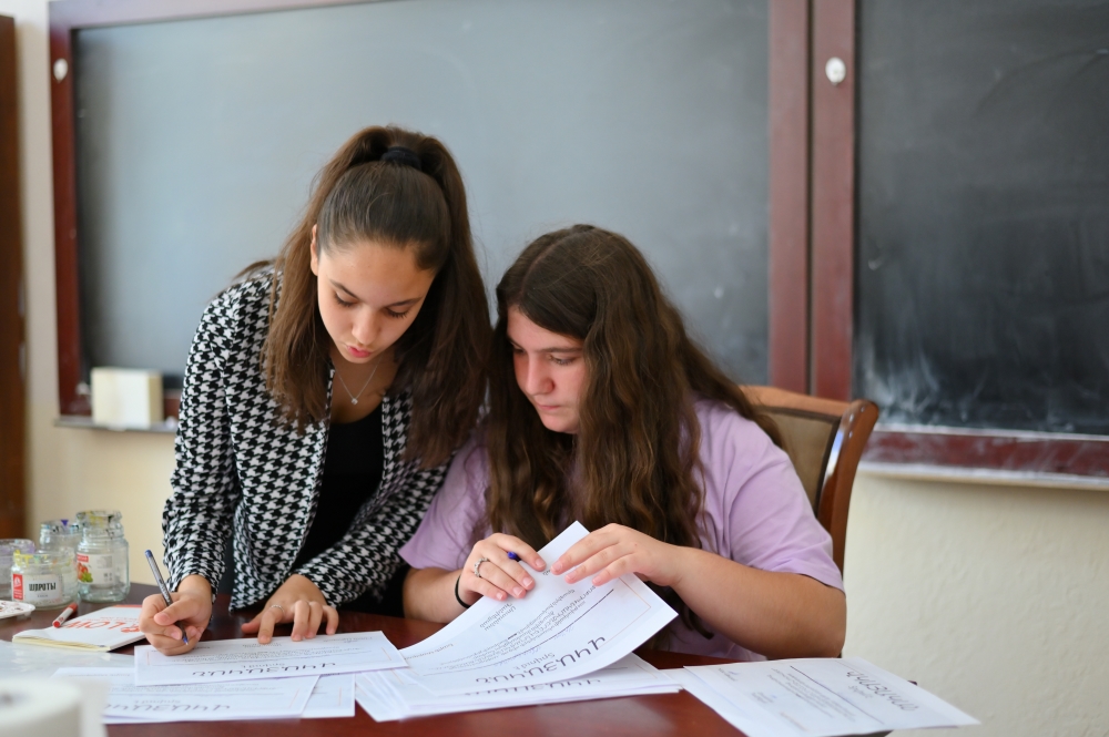 Two girls collaborating over paperwork at a table in a classroom with a blackboard in the background.