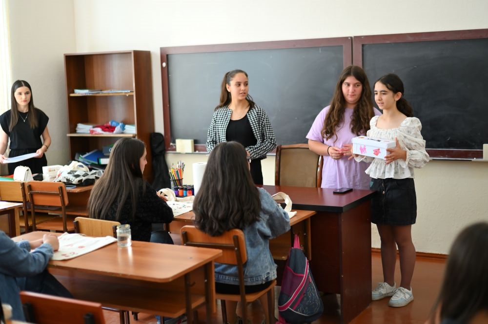 A classroom scene with several students standing and sitting around desks, interacting with each other. Two students at the front are giving a presentation.