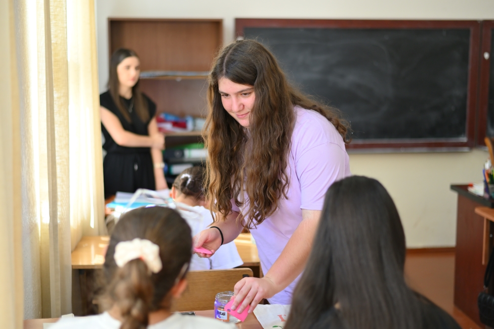 A girl handing out small papers to students in a classroom with a young girl standing by the blackboard in the background.