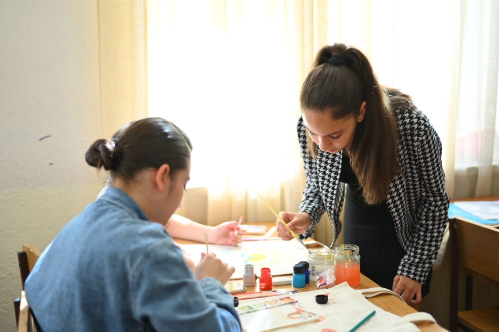 Two girls engaged in painting at a table, using various colors and materials. Bright sunlight streams through a window, illuminating the room.