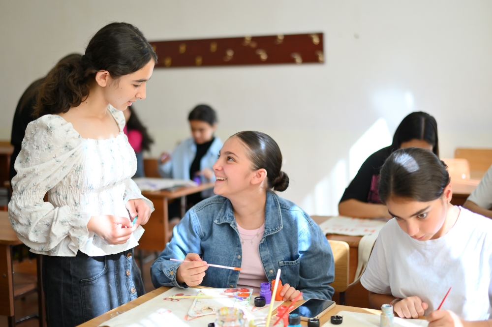 Two students engage in a conversation while working on an art project in a classroom setting, with another student focused on painting in the foreground