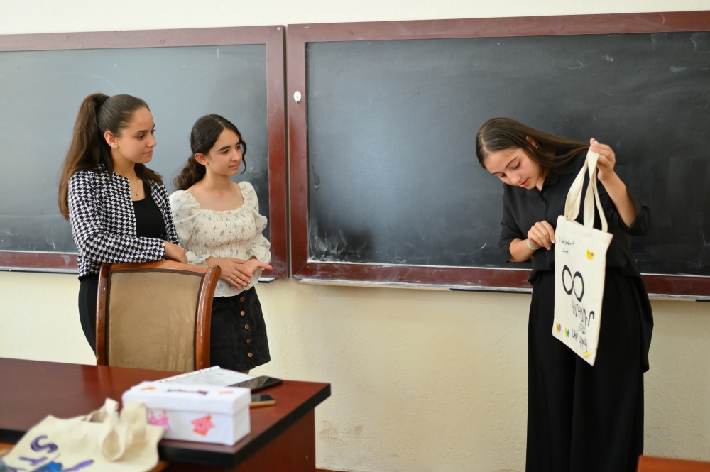 Three girls in a classroom setting, with one person showing her tote bag while the other two watch attentively at her. 