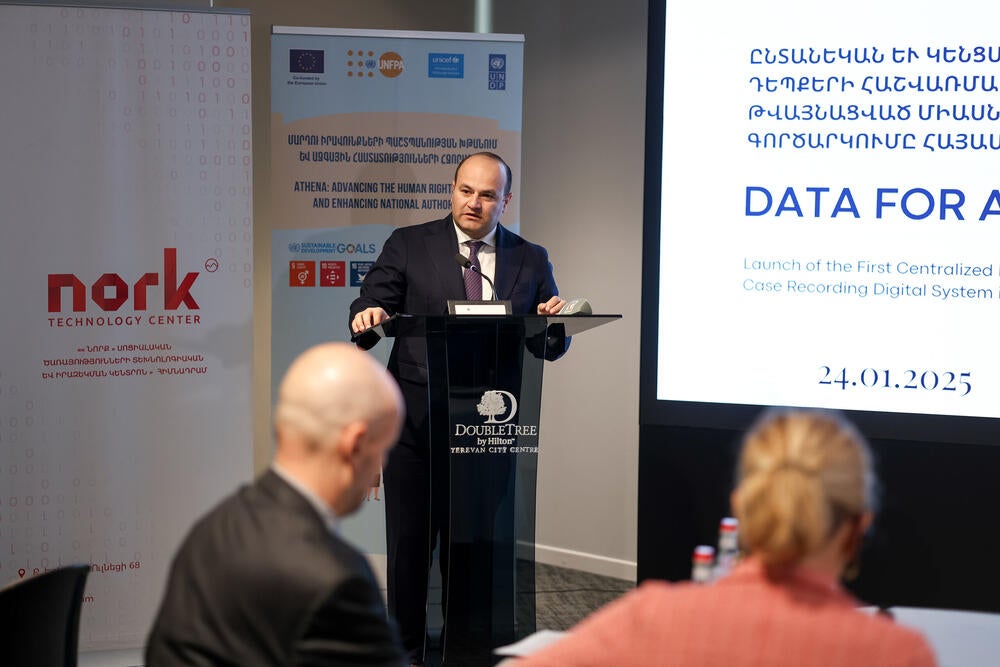 A person is presenting at a podium with a microphone at a 'DATA FOR ALL' event, with banners displaying logos including Nork Technology Center, EU and UNFPA. The audience, seen from behind, is listening attentively.