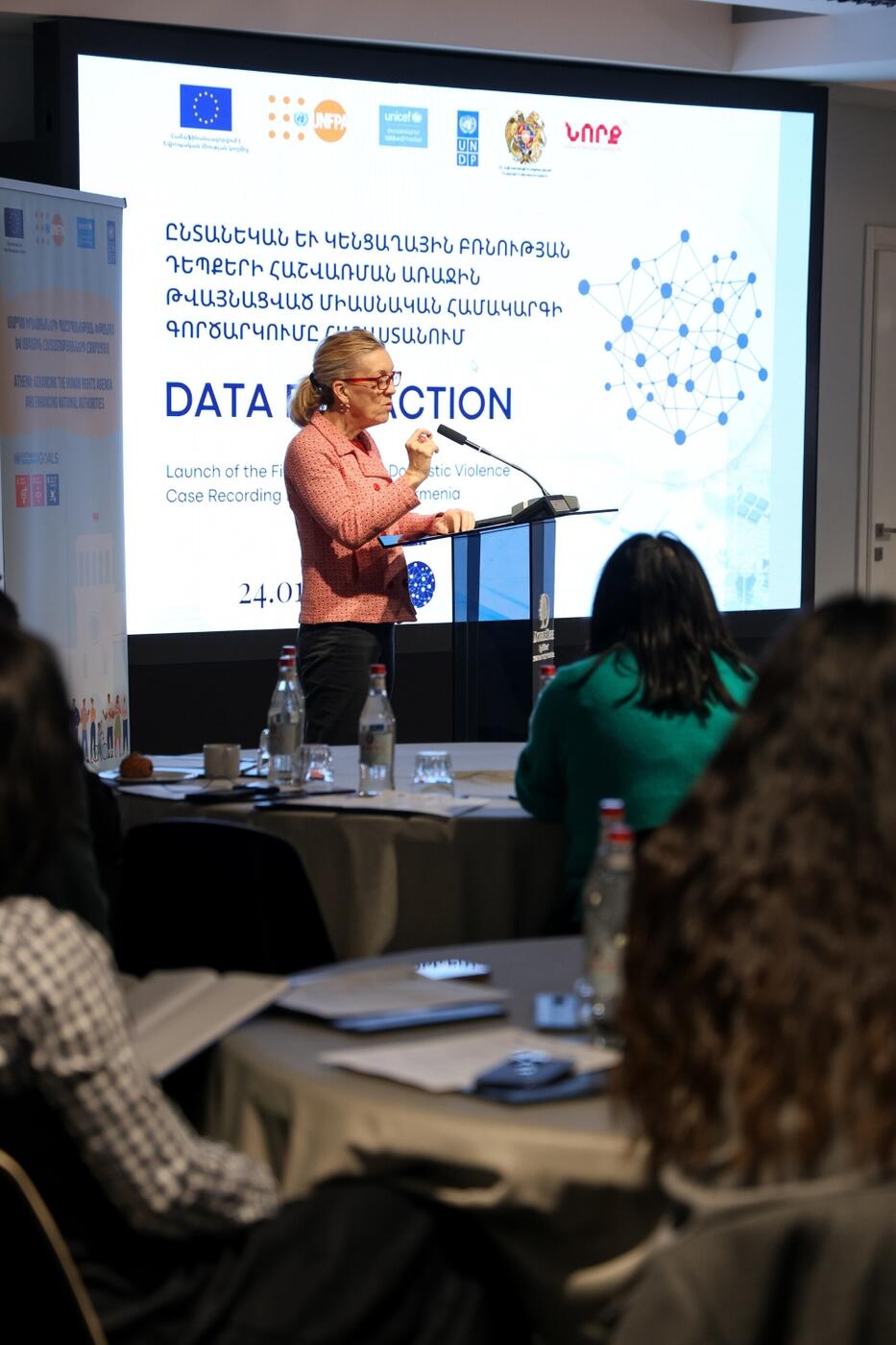 A person speaks at a podium during a conference on data on GBV, with a presentation screen displaying a graph and text in the background and an audience of attendees in the foreground.