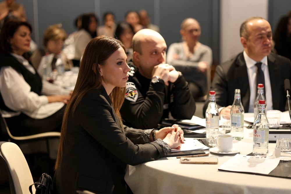 Audience members listening attentively at a conference, including a person in uniform and individuals in business attire. There are water bottles and notepads on the table in front of them.