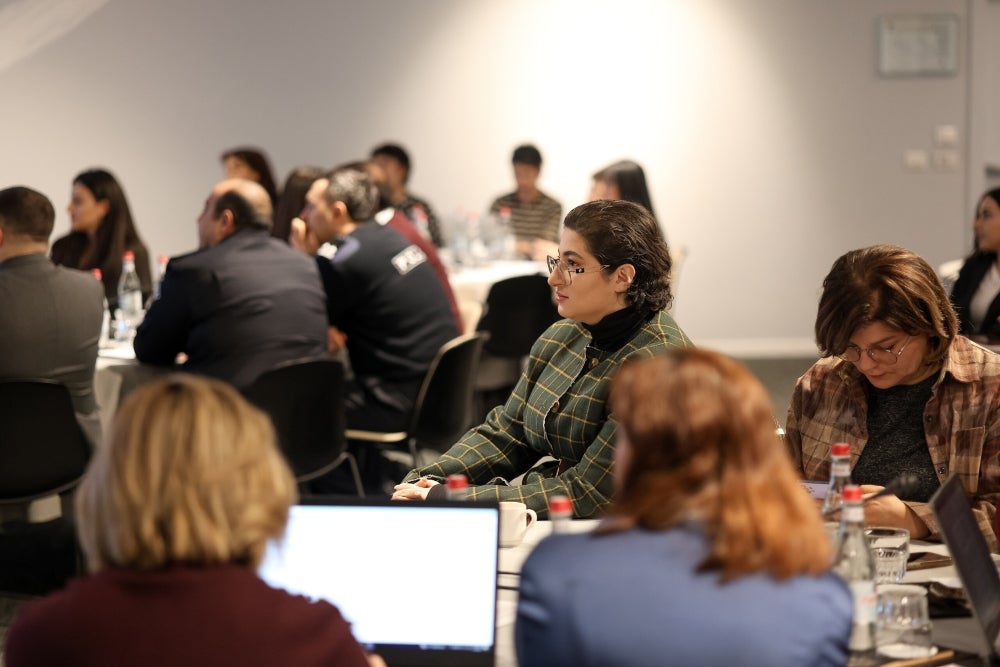 Attendees seated at tables during a conference session, focusing on presentations and taking notes. The setting includes laptops, notebooks, and a bright, modern room.