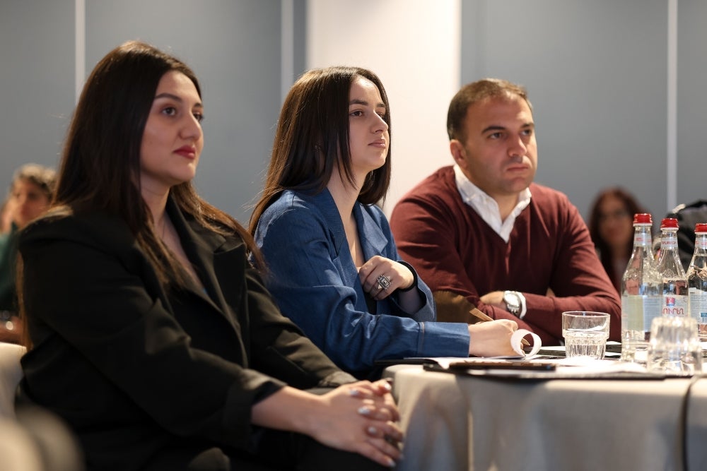 Three professionals attentively listening at a conference, seated in a row with water bottles and notepads on the table in front of them.