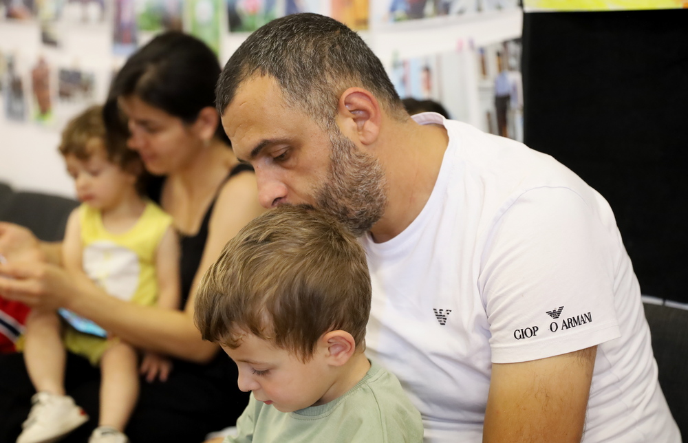 A father is holding his son in the foreground, with a mother and another child visible in the background.