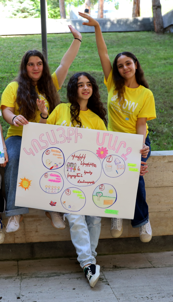 Three young girls in yellow shirts holding a large poster with various drawings and text. 