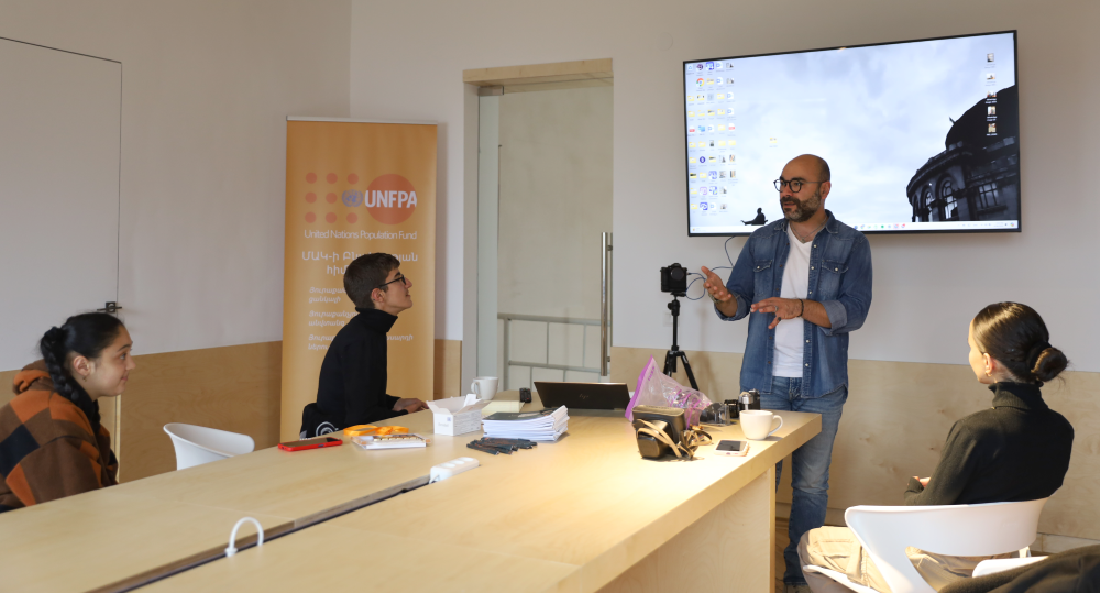 An individual presenting in a room with a large screen displaying a cityscape, to three young people listening him, near a banner of UNFPA (United Nations Population Fund).