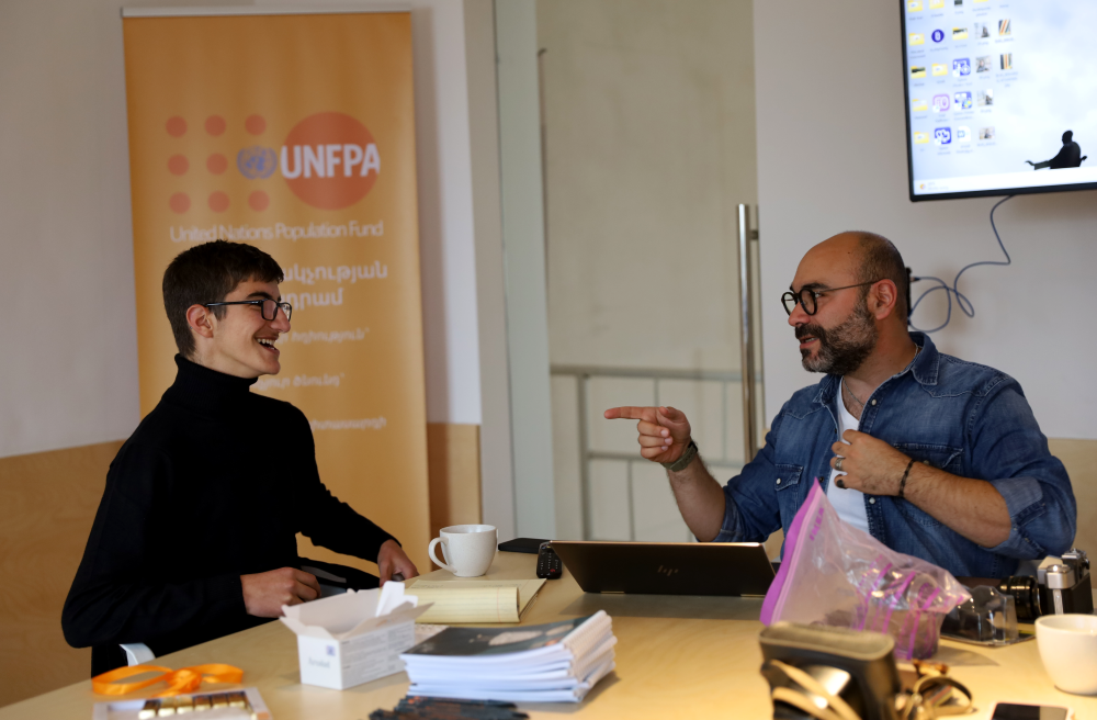 Two people engaging in a friendly conversation at a table with laptops and documents, in a room with a UNFPA (United Nations Population Fund) banner in the background.