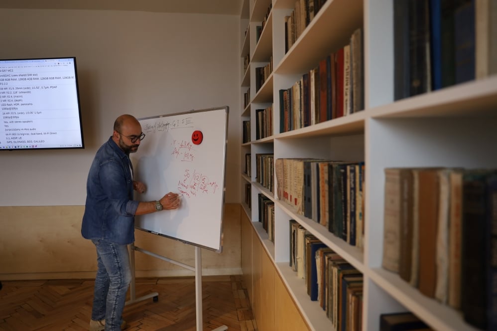 A man is writing on a whiteboard in a library surrounded by bookshelves, with a presentation being displayed on a monitor in the background.