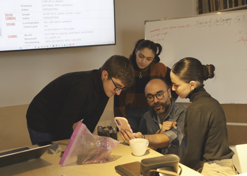 Four people collaboratively looking at a smartphone in a workshop: A young man was holding the smartphone and explaining something, the tree other adults: two girls and a boy are attentively looking at the phone. In the background featuring tech-related notes and diagrams on a whiteboard.