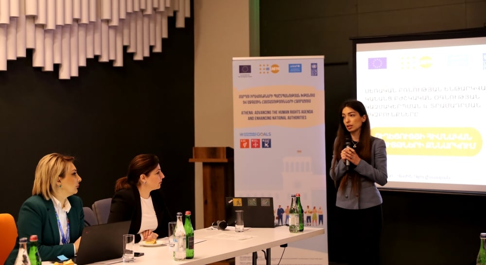 A woman presenting at a conference with two listeners seated at a table, beside banners displaying logos and text in a foreign language.